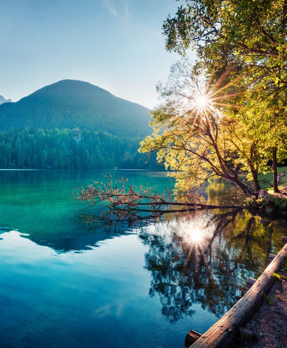 A lake with a mountain in the background reflecting off the water, the sun peeking through the trees.