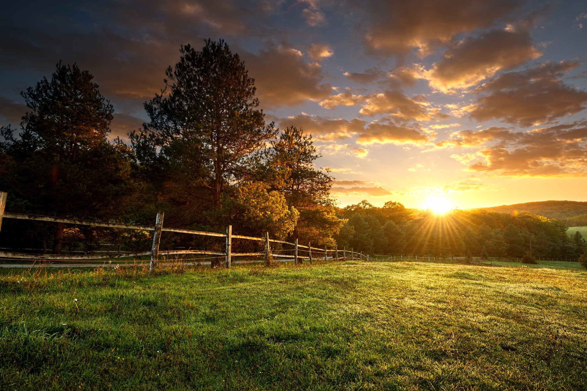 A golden sunrise cast light over a peaceful countryside landscape. The sun peeks through scattered clouds, illuminating a green field enclosed by a rustic wooden fence.